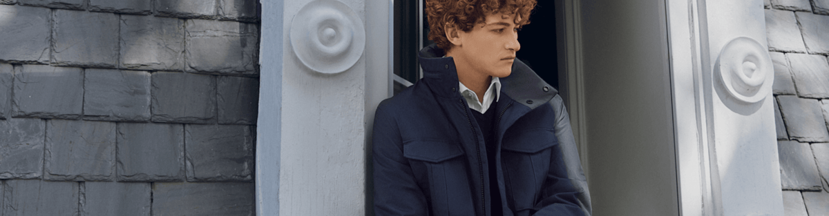 A man wearing a dark navy blue coat stands in a doorway with gray shingled walls on either side.